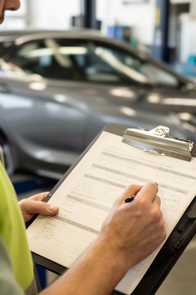 Technician writing vehicle inspection notes beside a parked sedan